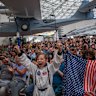A young boy wears an astronaut costume next to a woman waving a flag as they watch a live broadcast of the return of the Artemis II crew.