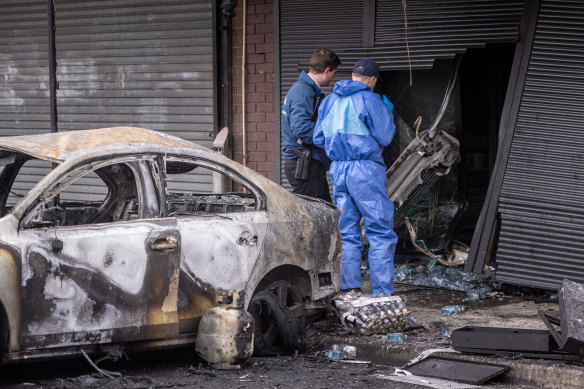Detectives collect evidence from the scene where a car was rammed into a greengrocer and set on fire.