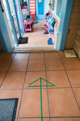 At Florence Rd. Pre School, tape on the ground indicates where parents and children should queue. A teacher lets students in one by one. 