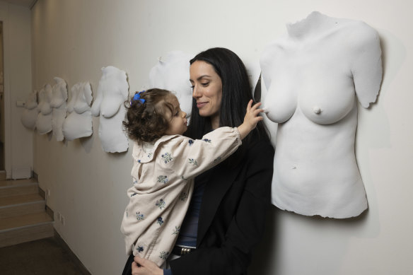 Amelie Cazzulino’s breastfeeding daughter, Gabrielle reaches towards a cast of her mother in the exhibition The Wall of Wombs by artist Juliana Monteiro.