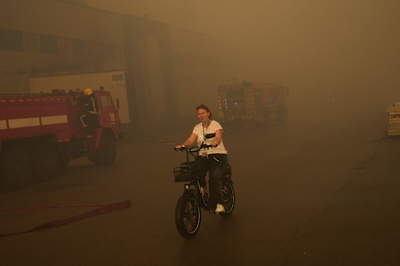 A woman rides an e-bike as firefighters work to extinguish a burning pharmaceutical warehouse attacked by Russia.