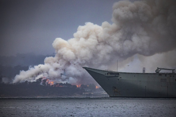 Fires burning on the southern side of Twofold Bay in Eden on Monday, with the bow of HMAS Adelaide anchored nearby.