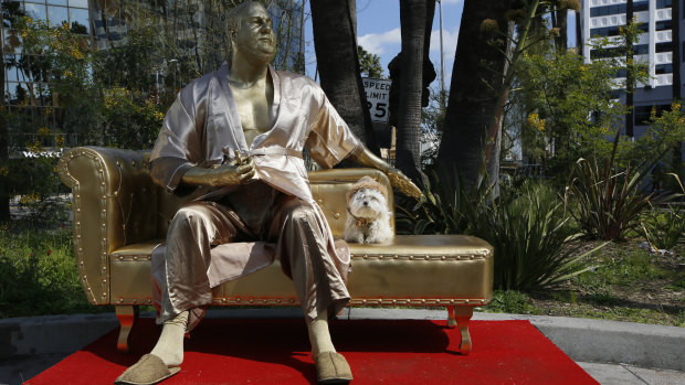 A dog sits next to the golden statue of a bathrobe-clad Harvey Weinstein.