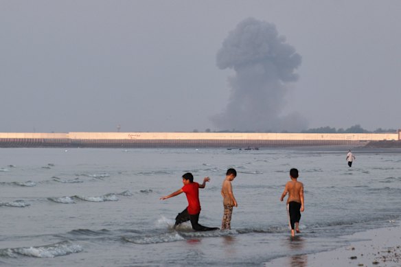 Three children play in the shallow waters of the Strait of Hormuz as smoke rises from an explosion in the background on Monday.