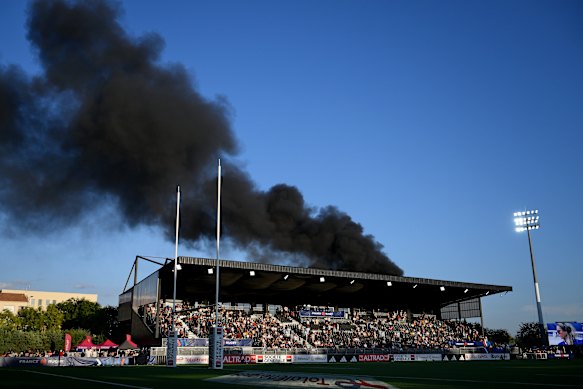 A cloud of black smoke billows out of a burning shuttle bus and drifts across the field during the WXV Global Series match between France and Australia.