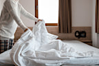 Woman making bed in bright bedroom. Maid Cleaning messy hotel bed cushion and duvet, changing bed sheets with clean. Selective focus. Copy space