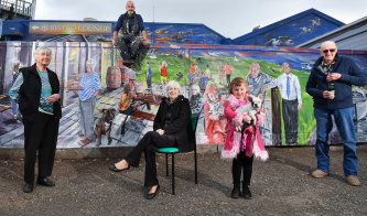 Artist Simon White, sitting on fence, with his mural on the LakeView Bar and Bistro  in Loch Sport, depicting locals Noeleen Brown, left, Dulcie Benham, Renee Meers, 6, holding the publican's dog Pushka, and Donny Morton. 