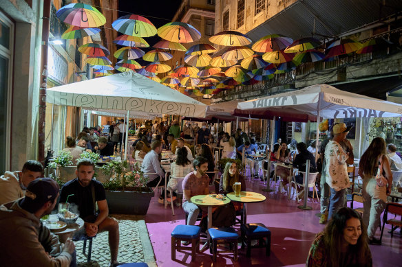 Tourists and locals sit at sidewalk tables under rainbow umbrellas in Pink Street, Cais do Sodre, Lisbon.