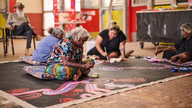 On the canvas, from left: Tuppy Goodwin, Puna Yanima, Sally Scales and Iluwanti Ken collaborate at Mimili Maku Arts.
Yaritji Young’s husband Frank sits on the chair.