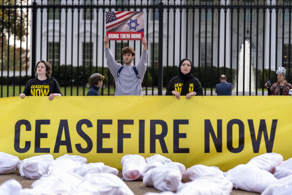 A man visiting from New Jersey holds up a poster in support of bringing Israeli hostages home behind a protest where a large banner that says “Biden: Ceasefire Now” along with fake white body bags.