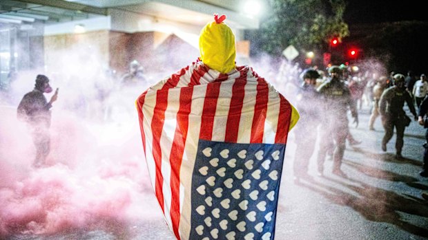 A protester clad in a chicken suit and an American flag with hearts instead of stars watches as law enforcement officers try to disperse protesters near a U.S. Immigration and Customs Enforcement facility in Portland.