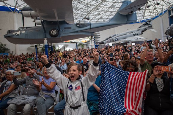 A young boy wears an astronaut costume next to a woman waving a flag as they watch a live broadcast of the return of the Artemis II crew.