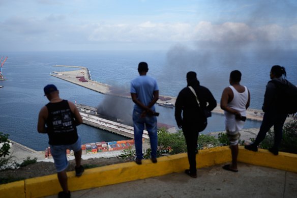 Men watch smoke rising from a dock after Venezuela’s La Guaira port was targeted by US forces.