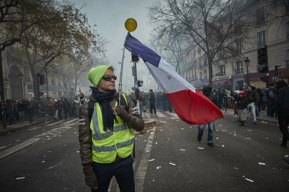 A <i>Gilet Jaune</i>, or Yellow Vest, holds a French flag as protestors and French riot police clash in Paris in 2019.