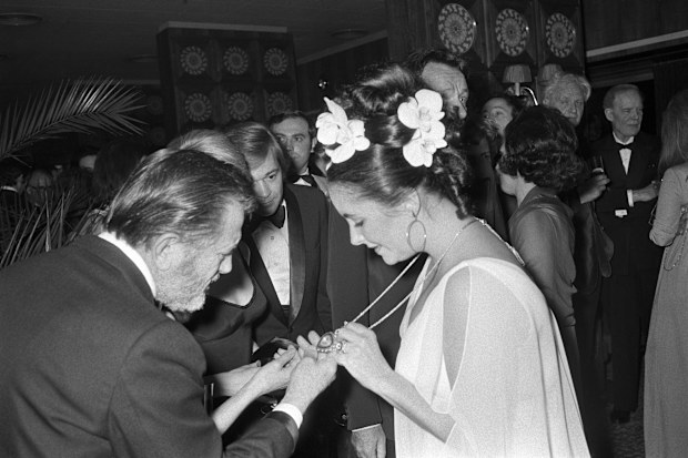 Guests at Elizabeth Taylor's fortieth birthday celebration brunch admire the actress' "Taj Mahal Diamond" pendant. The necklace, given to her by husband Richard Burton, is a lemon diamond inscribed with Indian love poetry suspended on a gold and ruby chain by Cartier. Taylor wears a white crepe chiffon Grecian dress designed by Edith Head with seven rare white orchids adorning her hair, designed by Alexandre of Paris. (Photo by Reginald Gray/WWD/Penske Media via Getty Images)