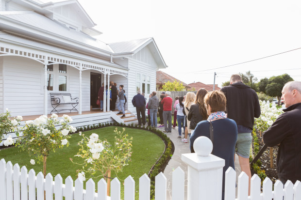 A line of people wait to be let into a white weatherboard property with a white picket fence and manicured garden.