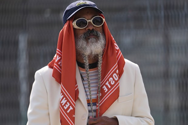 Juares Tenorio wears a blue cotton baseball cap with a gray / orange / white striped print pattern knit sweater from Lacoste, an orange and white print pattern silk scarf scarf from Lacoste