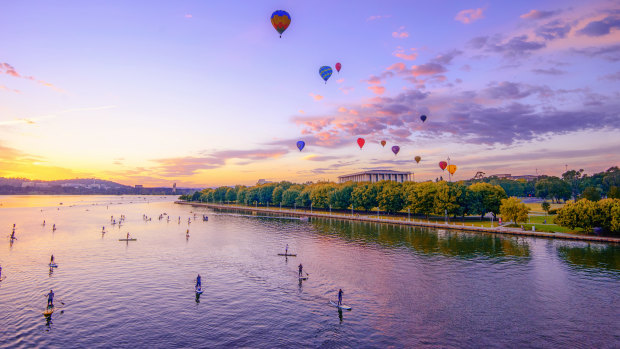 Balloons above the National Library as the sun rises over Lake Burley Griffin while paddle boarders take to the water.