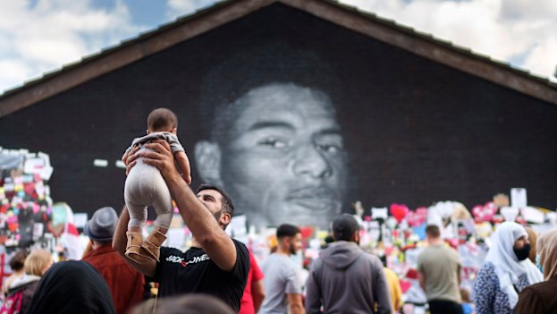 A man holds a child in the air as crowds gather at the Marcus Rashford mural in Manchester.