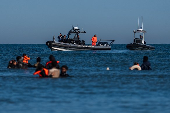 French police boats look on as migrants wade into the sea to board inflatables that they hope will carry them to England.