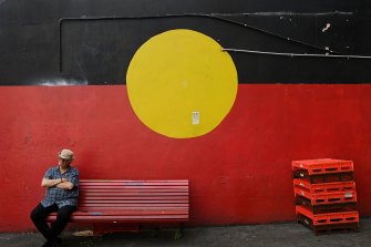 A man sits on a bench in front of a mural of the Aboriginal flag in Newtown, NSW.