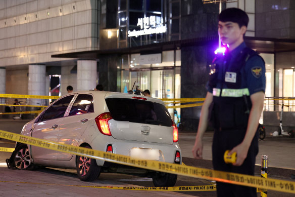 A white Kia hatchback with a broken front window and ruptured front tire on a sidewalk near a subway station.