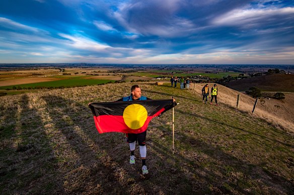 Travis Lovett unfurls the Aboriginal flag at the top of Mount Noorat during his Yoorrook Justice Commission Walk for Truth. The site was an important meeting place for First Nations people of western Victoria. 