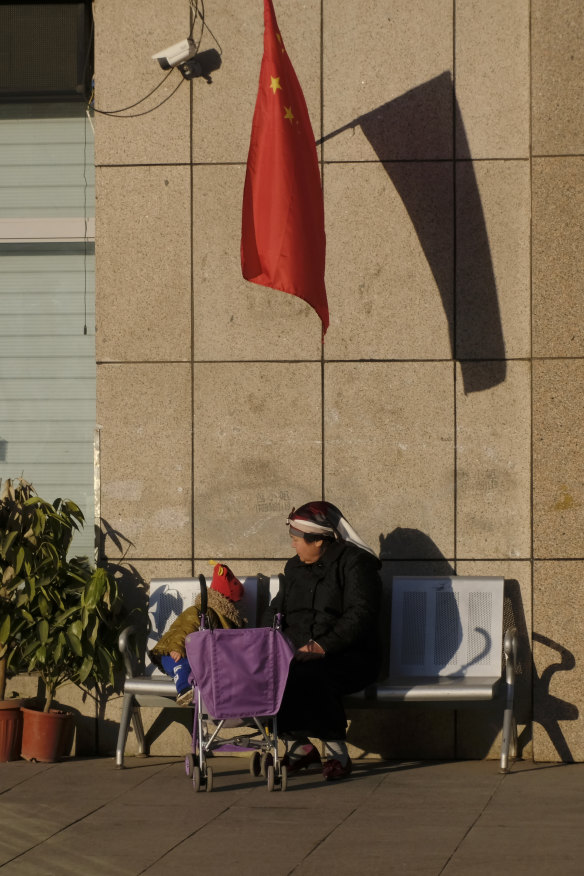 A Uighur woman and a child sit under China's national flag and a CCTV camera in Urumqi,  Xinjiang.