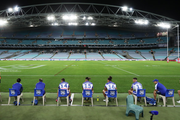 Bulldogs interchange players and support staff, seated well apart, look on at an empty ANZ Stadium.