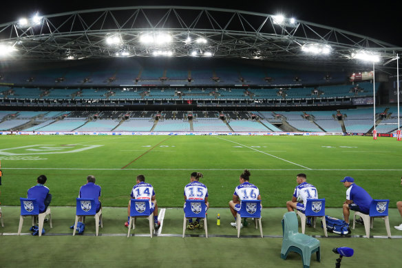 Bulldogs interchange players and support staff, seated well apart, look on at an empty ANZ Stadium.