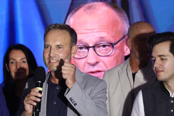 AfD members, including candidate Enrico Schult (second from left), speak to supporters at the party’s election evening gathering as Merz appears on a TV screen behind.