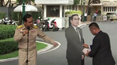 In this image from a video, Thai PM Prayuth Chan-ocha, left, directs the scene as a life-sized cardboard cut-out figure of himself is carried into view by an aide.