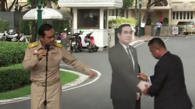 In this image from a video, Thai PM Prayuth Chan-ocha, left, directs the scene as a life-sized cardboard cut-out figure of himself is carried into view by an aide.