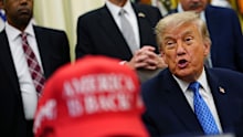 President Donald Trump speaks alongside a red cap stating "America Is Back!" in the Oval Office of the White House.