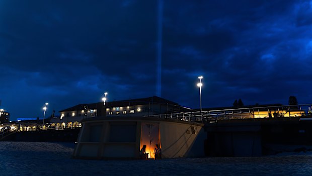 People light candles on Bondi Beach while light beams are projected from Bondi Pavilion to mark seven days since a terrorist attack killed 15 people.