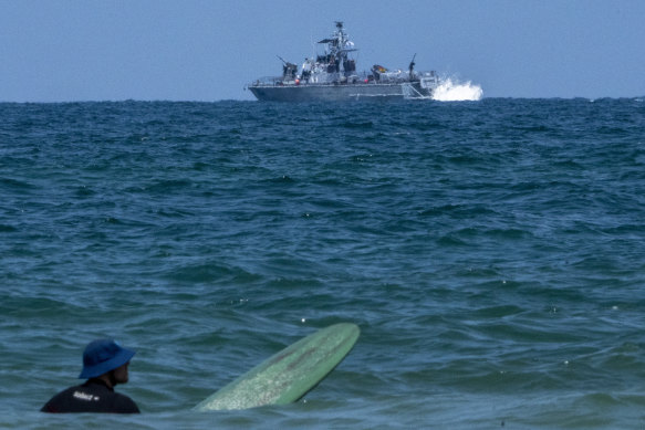A surfer waits for wave while an Israeli military naval ship patrols the Mediterranean sea off the coast of Hadera, Israel.