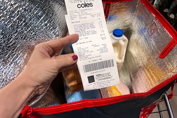 A Coles receipt being held up in front of a trolley containing a bag of groceries.