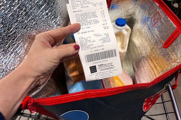 A Coles receipt being held up in front of a trolley containing a bag of groceries.