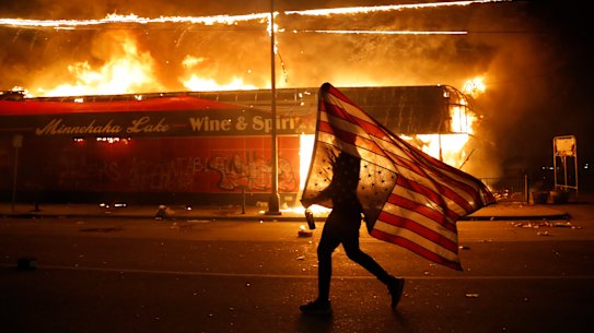 A protester carries the carries a U.S. flag upside, a sign of distress, next to a burning building Thursday, May 28, 2020, in Minneapolis.