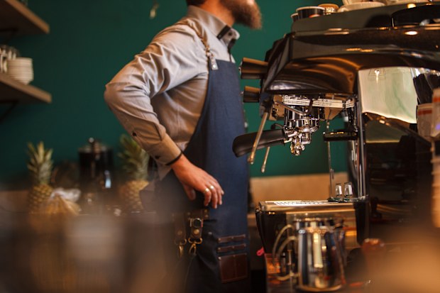 Side view of unrecognizable barista standing behind bar counter, hands on hips, taking customer's order, espresso machine ready to go.