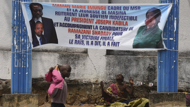 A child street beggar walks past a woman selling medicine on a street in Kinshasa under a banner supporting outgoing President Joseph Kabila and his hand-picked replacement, Emmanuel Ramazani Shadary.