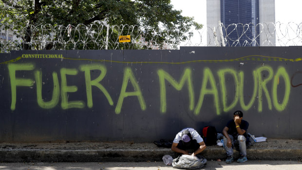 A man reaches into his bag to retrieve some food waste that was given to him by a Chinese restaurant as he sits in front of security fence spray-painted with a message that reads in Spanish: "Maduro out," in Caracas, Venezuela.