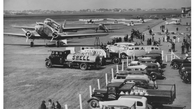 Aircraft and petrol tankers outside the hangers at Essendon Aerodrome. People picnic on the grass or stand around chatting while waiting to board, 1937.