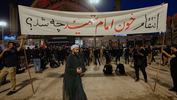 A cleric walks past mourners carrying a banner reading “What is the revenge for the martyrdom of our Imam?”