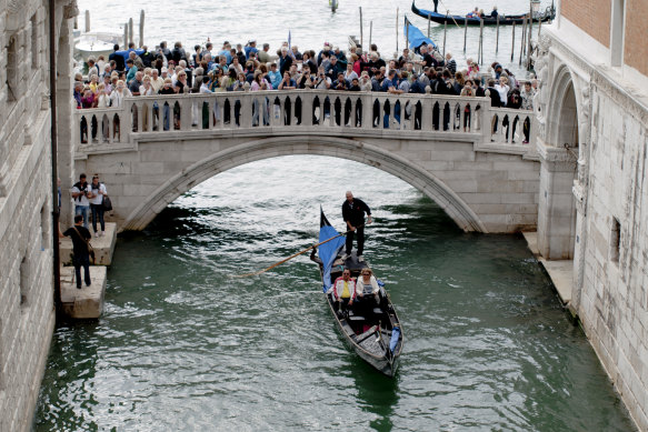 Traditional gondola carrying passengers pass under a bridge full of tourists near Piazza San Marco in Venice.