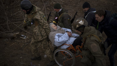 Ukrainian soldiers and militiamen carry a woman in a wheelchair as the artillery echoes nearby while people flee Irpin.