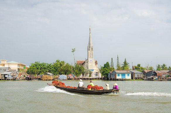 Estrechos barcos de madera cargados de frutas y verduras cruzan el Mekong, con la iglesia de Cai Be al fondo.