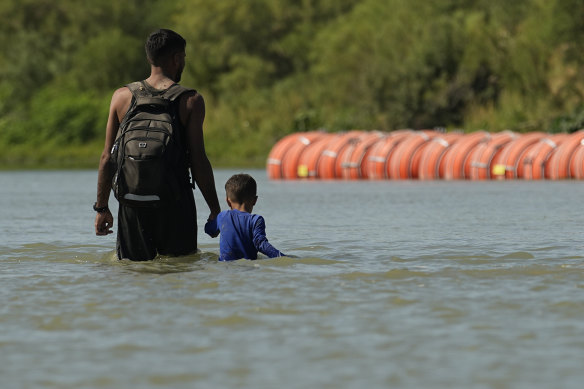 Migrants walk past large buoys being used as a floating border barrier on the Rio Grande River between Texas and Mexico.