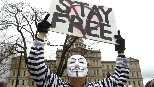 A protester wearing a Guy Fawkes mask holds up a sign in Lansing.