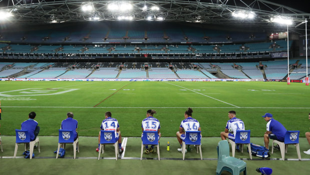 Bulldogs interchange players and support staff, seated well apart, look on at an empty ANZ Stadium in round two.